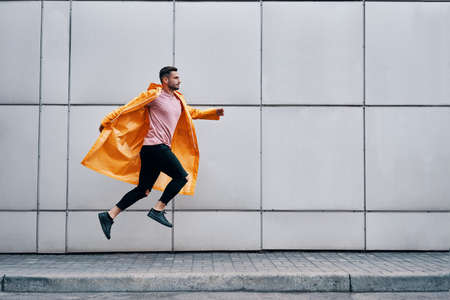 Trendy young man in yellow raincoat jumping in the air on wall street urban backgroundの写真素材