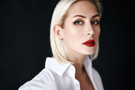 Close up portrait of beautiful blonde woman with red lips in elegant white shirt posing isolated on black studio backgroundの写真素材