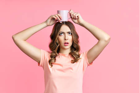 Young pretty woman having fun with cup of coffe on her head on pink background