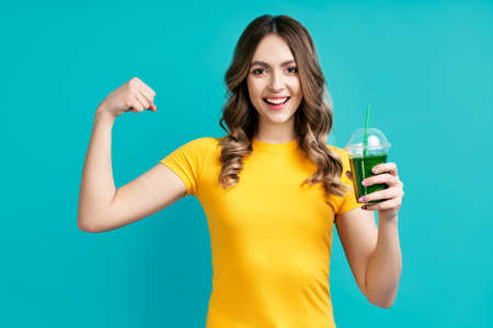 Pretty young woman holding green detox vegetable smoothie juice and shows muscles