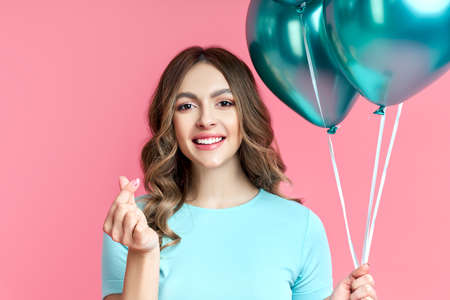 Pretty smiling woman makes mini heart korean love symbol by fingers and holding blue balloons on...