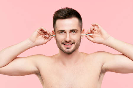 Young smiling man cleaning ears with cotton swab over pink backgroundの写真素材
