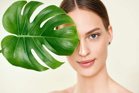 Close up portrait of beautiful young woman covering her eye with monstera tropical leafの写真素材