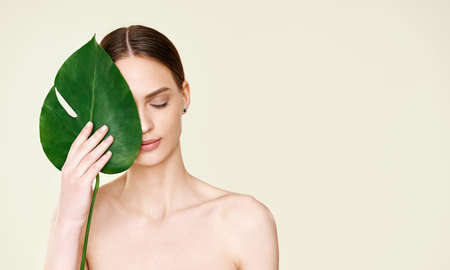 Close up portrait of young beautiful woman with perfect smooth skin covering her eye with monstera tropical leafの写真素材