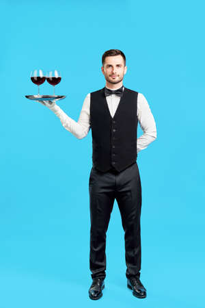 Full length portrait of elegant male waiter with glasses of wine over blue backgroundの写真素材