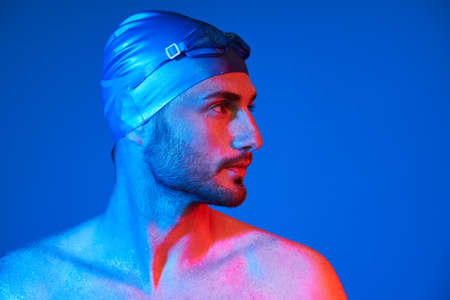 Close up profile portrait of handsome swimmer with goggles in red-pink neon light over blue background with copy spaceの写真素材