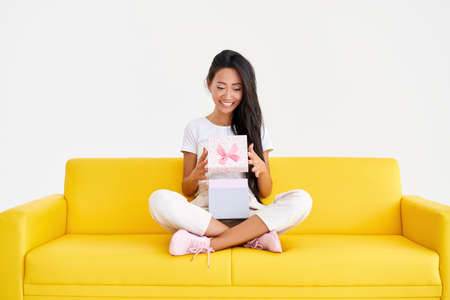Happy smiling woman opening a gift box sitting on comfortable sofaの写真素材