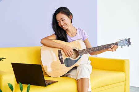 Smiling pretty female learning to play guitar watches a video lesson online using laptop