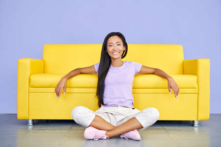 Happy smiling asian woman relax sitting on floor leaning yellow sofaの写真素材