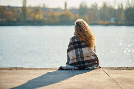 Back view of young woman relaxing alone by a river in the city wrapped in wool blanket enjoying natureの写真素材
