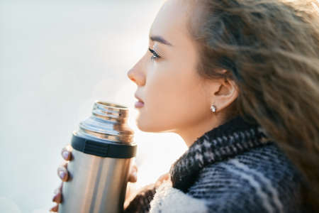 Young pretty woman  drinks tea wrapped in blanket over blur river background.の写真素材