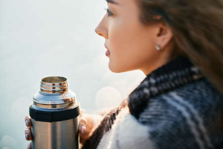 Young pretty woman drinks tea wrapped in blanket over blur river background.の写真素材