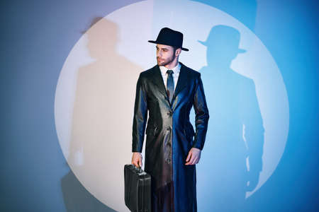 Handsome man in black coat and hat holding briefcase posing in the spotlight on studio backgroundの写真素材