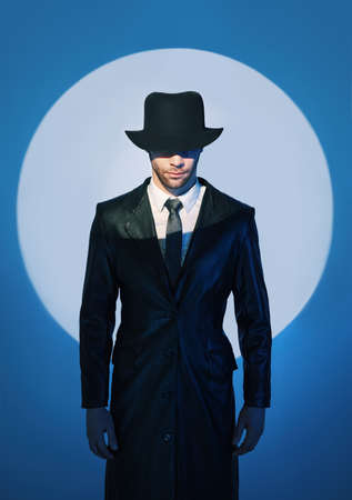 Portrait of handsome man in black coat hiding face behind his hat in the spotlight on studio backgroundの写真素材