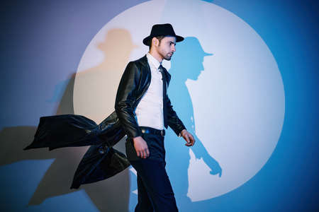 Handsome man in black coat and hat walking like superhero in the spotlight