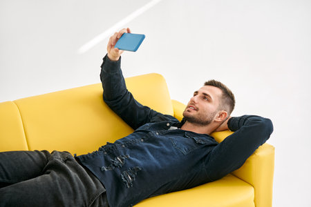 Relaxed man lying on yellow sofa talk on video call by his mobile phone over white background
