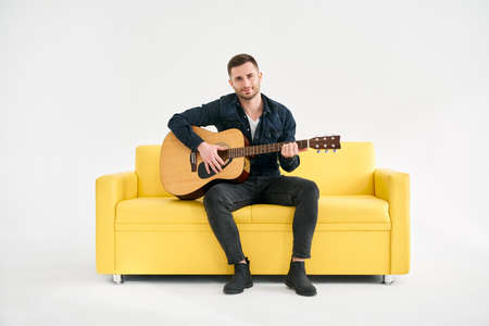 Handsome young man playing acoustic guitar whlile sitting on yellow sofa over white backgroundの写真素材