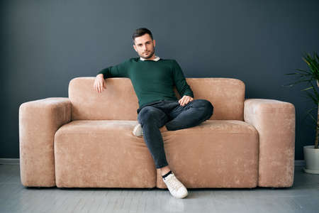 Handsome trendy man posing looking to camera while sitting on comfort sofa in modern apartmentの写真素材