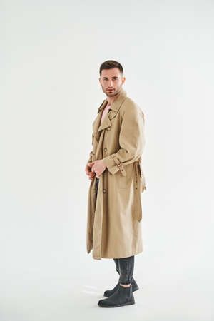 Full length portrait of young trendy man in beige trench coat over white backgroundの写真素材