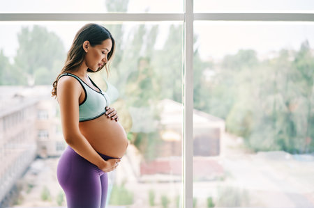 Portrait of young pregnant woman in sportswear standing near window holding hands on her belly with copy spaceの写真素材