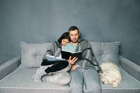 Smiling young couple reading books together on couch under plaid at home