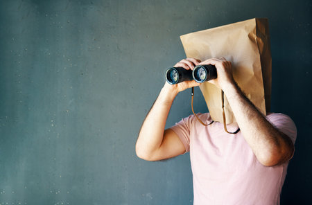Man with a paper bag on head looking through binoculars to copy space