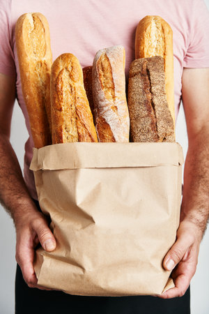 Man holds fresh loaves of bread baguettes in paper bag over light backgroundの写真素材