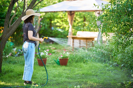 Young woman in hat watering flowers and plants in garden with hose in sunny blooming backyard
