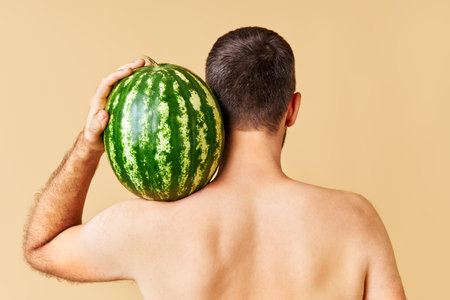 Back view of young man holding fresh watermelon on shoulderの写真素材