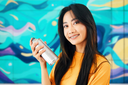 Portrait of smiling female street artist with spray paint can in hand looking to camera over her...