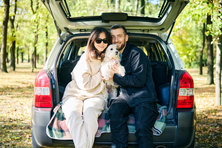 Loving couple hugging their fluffy white dog while relaxing in car trunk in forest. Family, vacation, holidays, travel conceptの写真素材