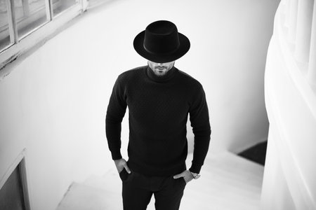 Stylish man in black clothing outfit and hat, looking downwards walking on marble staircase. Black and white photoの写真素材