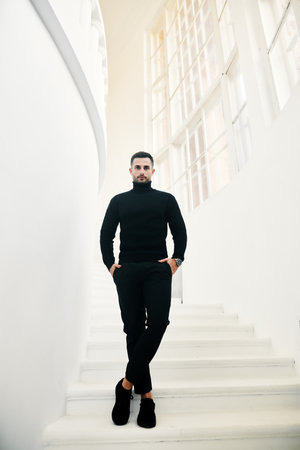 Handsome fashionable man model in an all-black clothing outfit posing on white staircase. Male beautyの写真素材