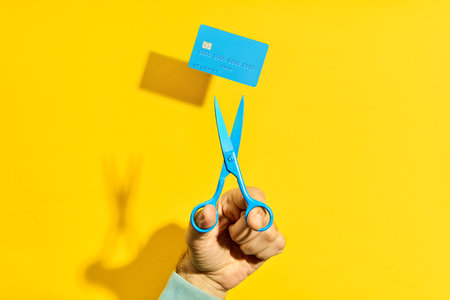 Hand holding blue scissors cutting blue credit card on bright yellow background. Finance concept