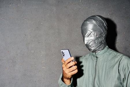 Mystery man with duct tape face types text message on cell phone, enjoys online communication