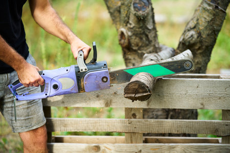 Close-up of man cutting trees using electrical chainsaw in motion at backyard homeの写真素材