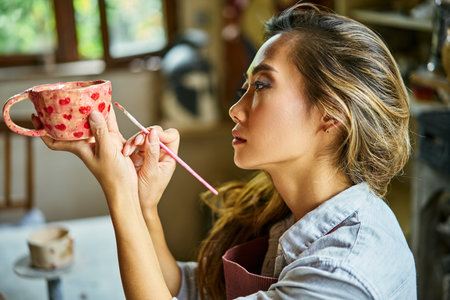 Female artist decorating and painting handmade ceramic clay cup at her creative workshop. Artwork and handmade conceptの写真素材