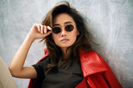 Fashionable confident woman wearing red coat, stylish casual outfit and sunglasses posing on gray background at studio.の写真素材