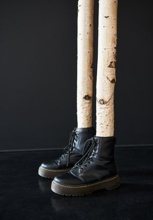 Creative surreal concept of birch tree trunks as human legs wearing black combat boots on dark...