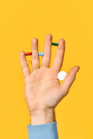Male hand holding various pills balanced on fingers over yellow background. Medicine, healthcare...