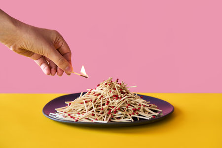 Hand holds burning matchstick above a large pile of matches on plate over vibrant background. Danger, risk conceptの写真素材