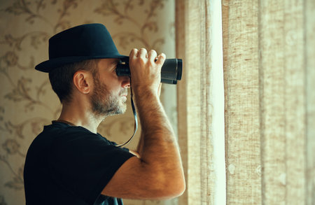 Man in fedora hat looking through binoculars spying from window. Spy and search conceptの写真素材