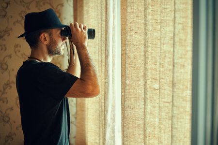 Man in fedora hat looking through binoculars spying from window. Spy and search concept