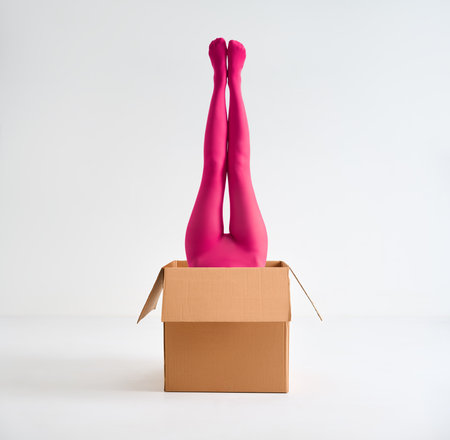 Female legs in bright pink tights raised up from open cardboard box over white studio backgroundの写真素材