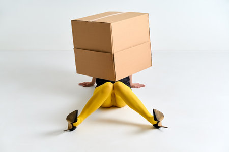 Woman in yellow tights and black high heels hidden under oversized cardboard box on white floor. Fun conceptの写真素材