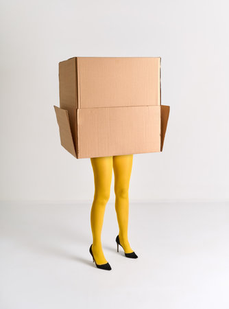 Woman in yellow tights and black high heels hidden under oversized cardboard box on white...