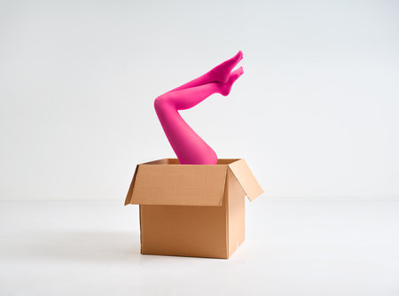 Female legs in bright pink tights raised up from open cardboard box over white studio backgroundの写真素材