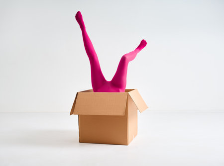 Female legs in bright pink tights sticking out of open cardboard box over white studio backgroundの写真素材