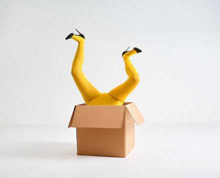Female legs in yellow tights and high heels shoes raised up in open cardboard box over white studio background. Fun conceptの写真素材