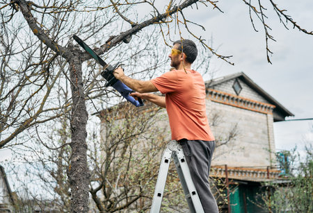 Young man cutting trees using electrical chainsaw in motion at his backyard. Gardening conceptの写真素材